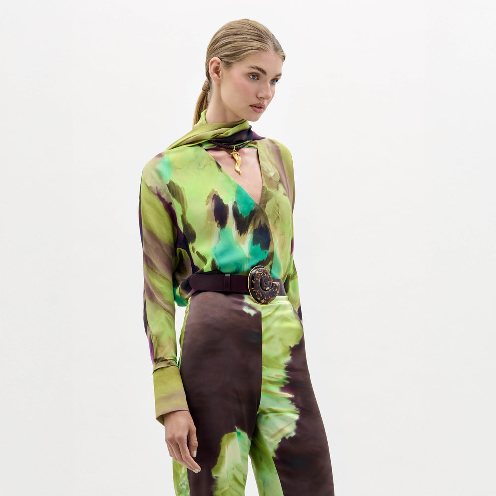 A woman stands against a plain white background wearing the Jaboca Blouse—a green, yellow, and brown tie-dye top styled with a wide belt and a scarf draped around her neck.