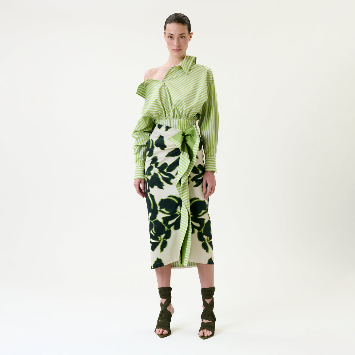 A woman stands against a white background wearing an asymmetrical green striped top, the Luana Skirt—a green and black floral wrap skirt—and olive green strappy heels.