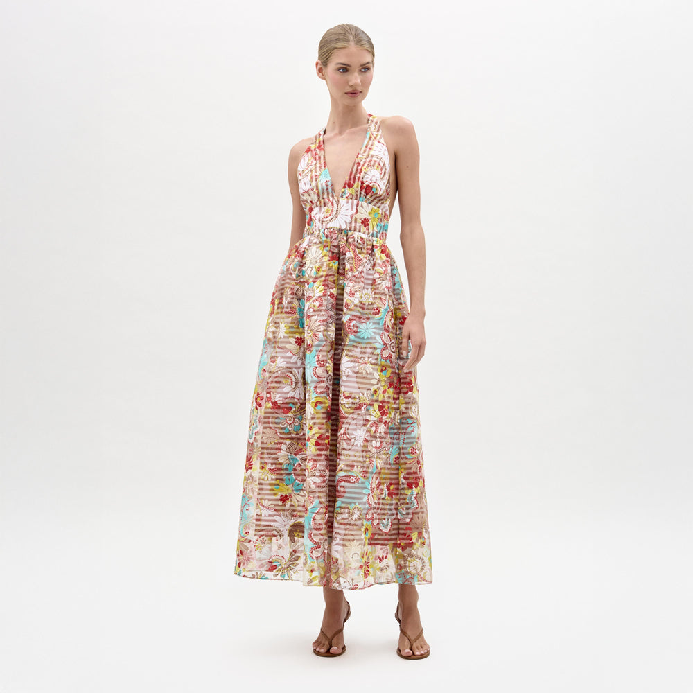 A woman models the Kairi Dress, a sleeveless, floor-length gown featuring a deep V-neckline and vibrant, intricate patterns on luxurious Italian fabric, standing against a plain white background.