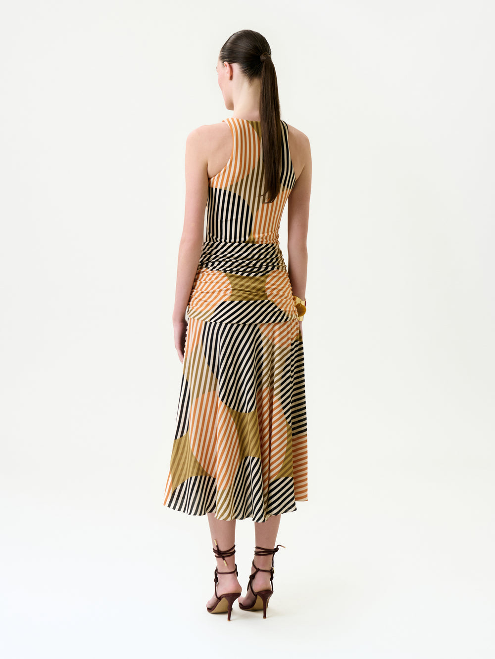 A woman with a long ponytail is standing, facing away, wearing the Malvina Dress—a sleeveless, calf-length midi crafted from luxurious Italian fabric with black, white, and tan geometric stripes—and brown strappy heels, all set against a plain white background.