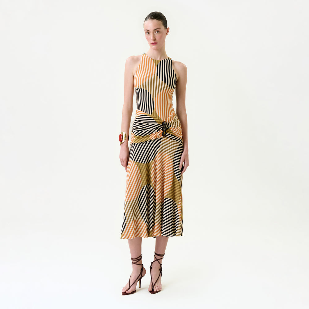 A woman poses against a plain white background wearing the Malvina Dress, a sleeveless, multicolored striped midi dress with a side knot crafted from luxurious Italian fabric, styled with strappy heels and a red cuff bracelet.