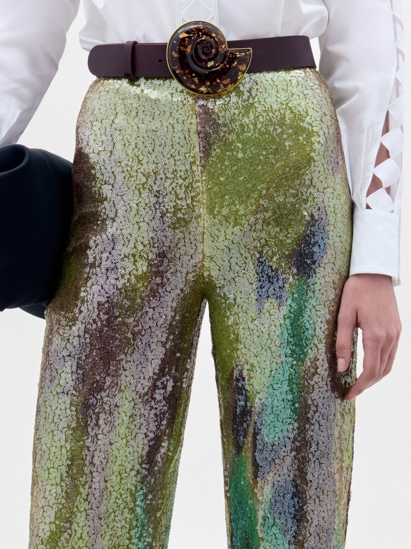 A person stands against a plain background wearing a white button-up shirt and the high-waisted, multicolored sequin Dalal Pant, holding a black clutch in their left hand.
