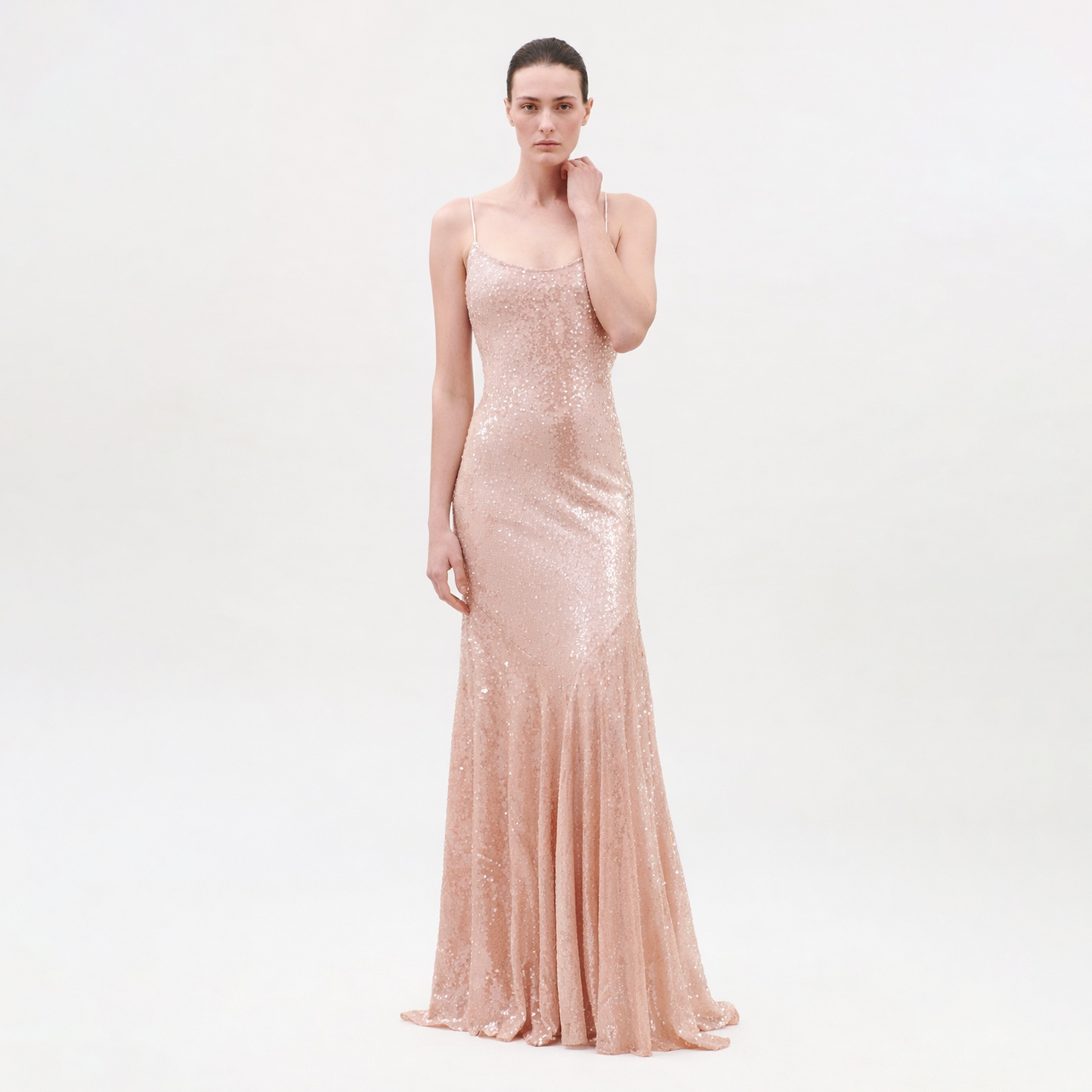 A woman with a neutral expression and pulled-back hair stands against a plain background wearing the Ibona Dress, a floor-length blush pink sequin gown with thin spaghetti straps and a form-fitting silhouette.