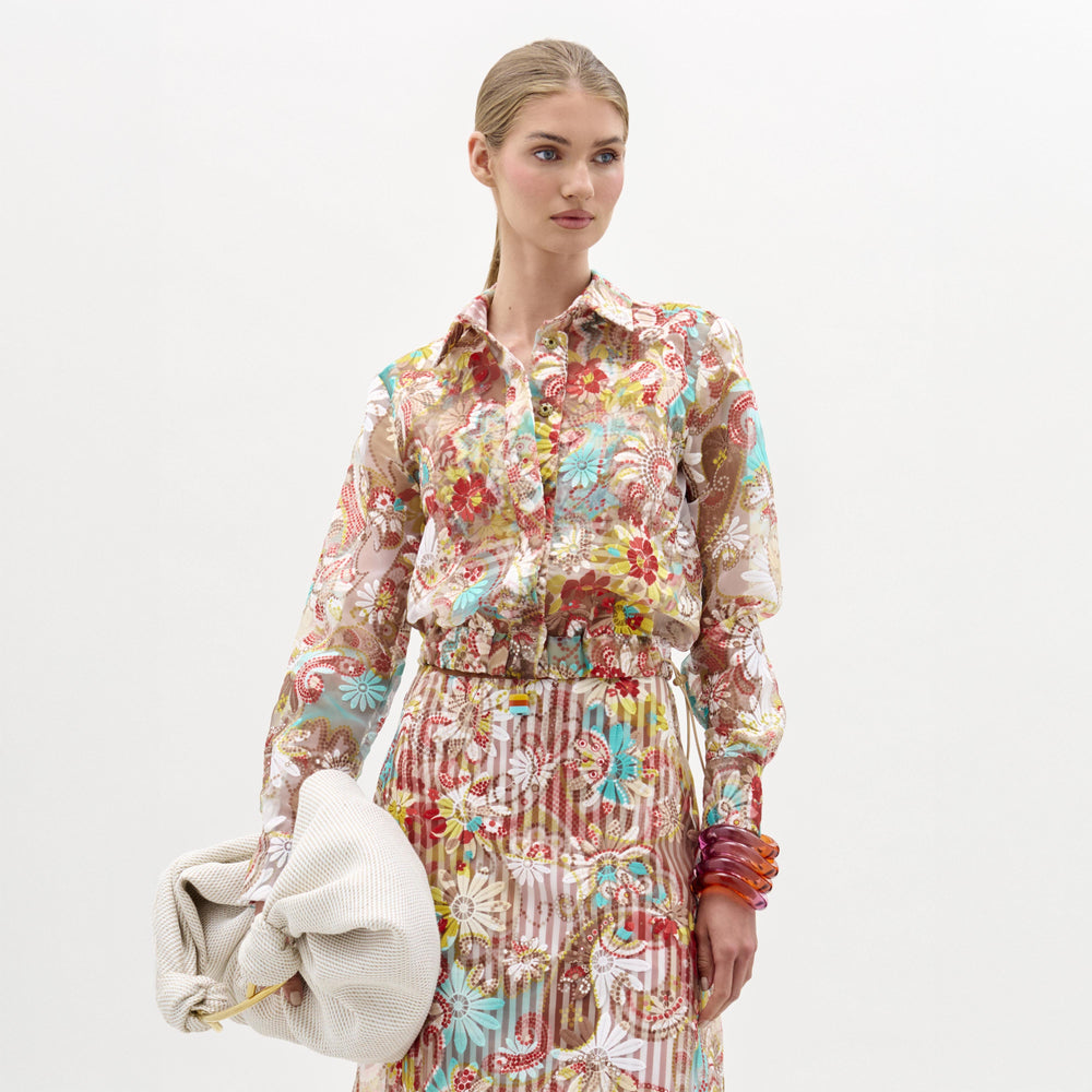 A woman wearing the Katniss Jacket crafted from Italian fabric, paired with a matching skirt, holds a large white textured clutch and stacked bracelets, standing against a plain background.