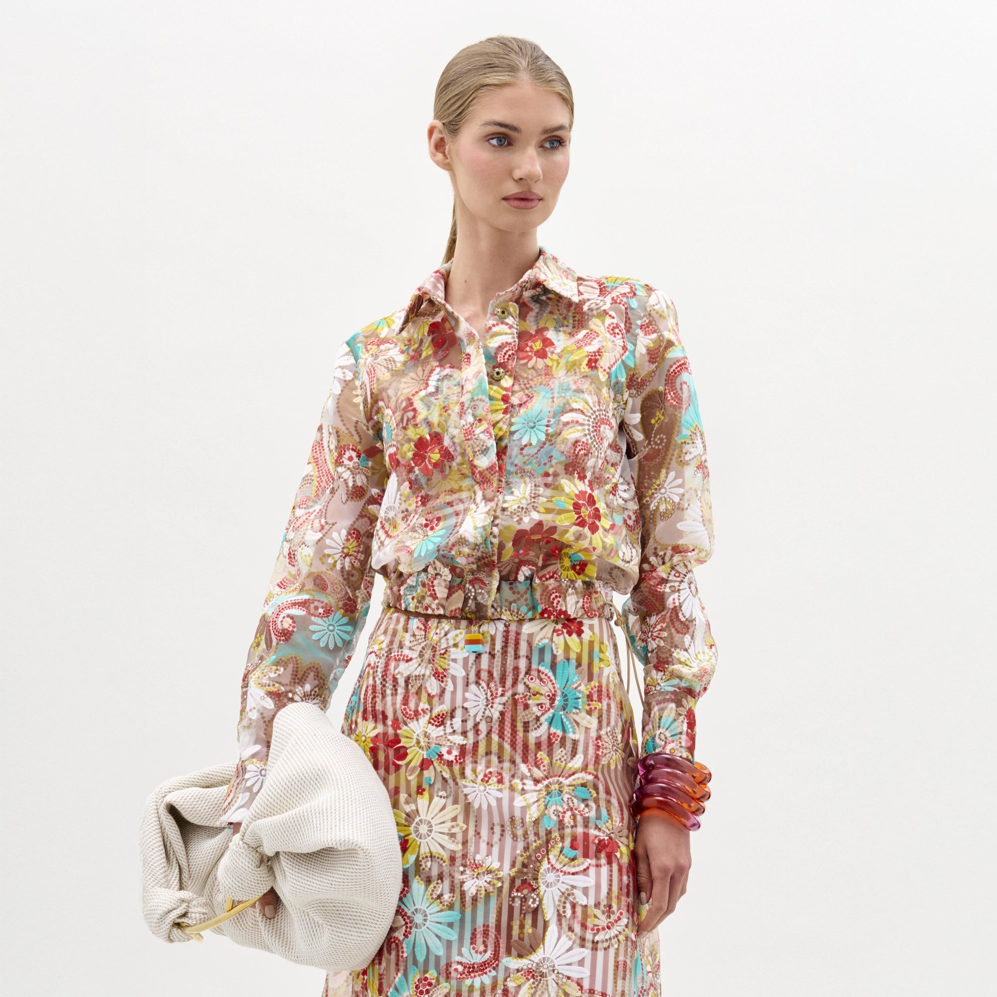 A woman wearing the Katniss Jacket crafted from Italian fabric, paired with a matching skirt, holds a large white textured clutch and stacked bracelets, standing against a plain background.