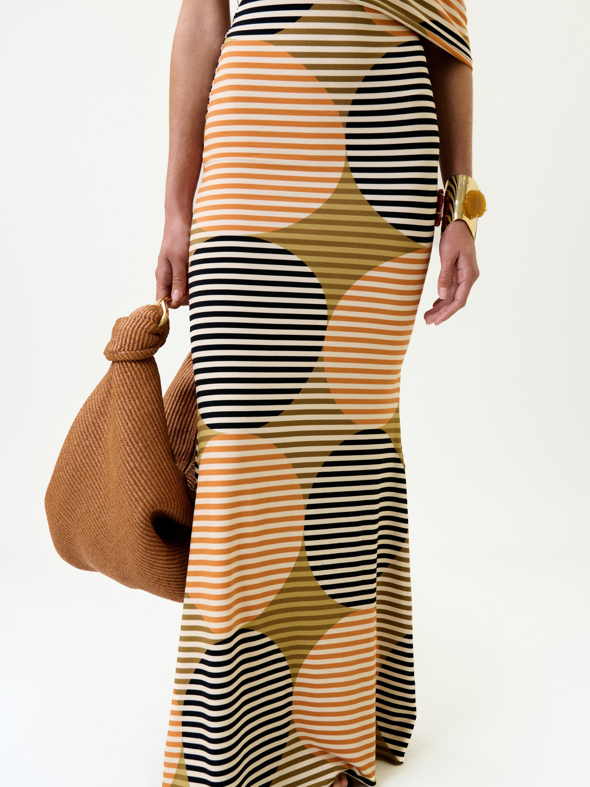 Wearing the Jensine Dress, made from Italian fabric with bold black, peach, and olive stripes and circular patterns, a person holds a textured brown bag and wears a chunky gold bracelet; their upper body is partly out of frame.