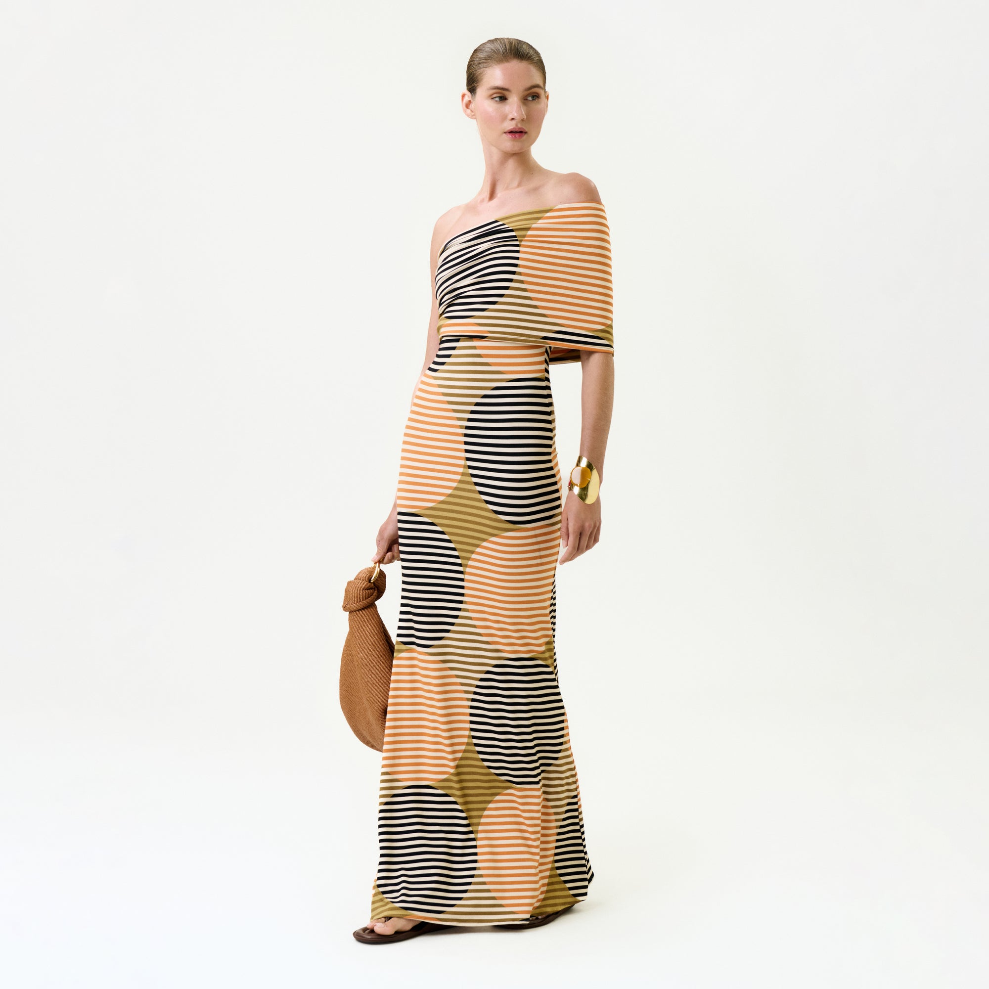 A woman poses against a plain background wearing the Jensine Dress, a one-shoulder fitted maxi made in Colombia with bold yellow, orange, black, and beige polka dots and stripes. She carries a brown bag and accessorizes with sandals and a chunky bracelet.