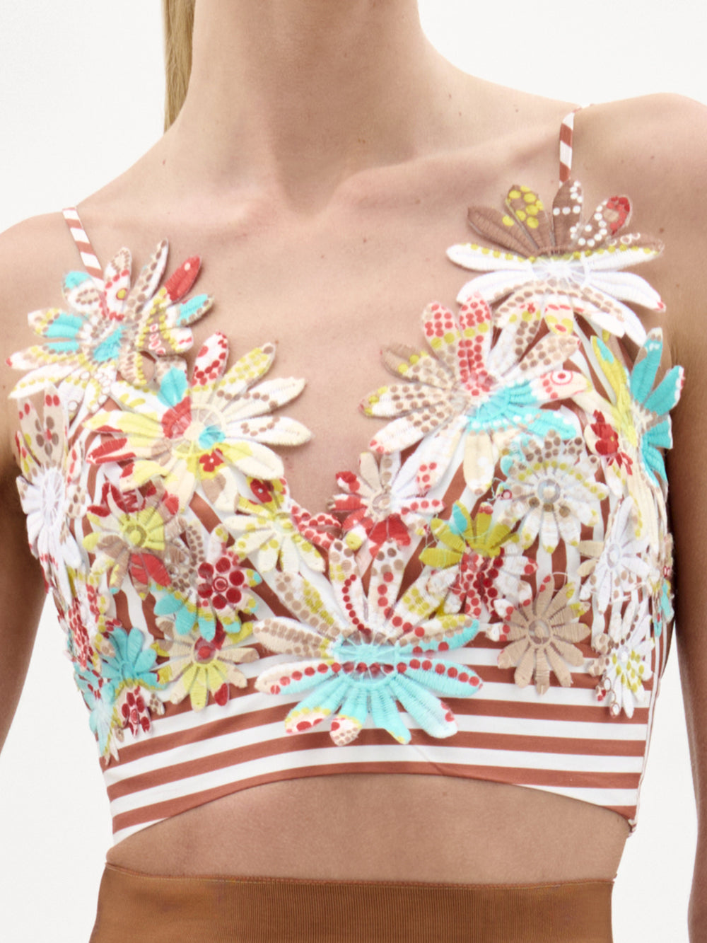 A woman wearing the Ibbie Top, a floral embroidered cropped top made from luxurious Italian fabric, pairs it with a matching skirt, holds a large textured clutch, and wears red bangles, standing against a plain white background.