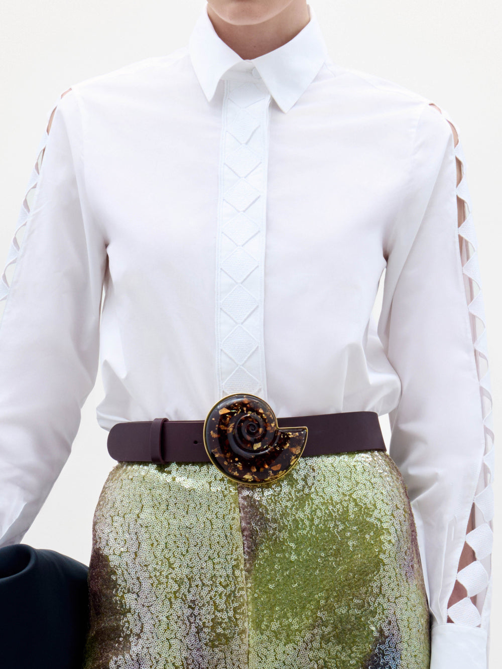 A woman stands against a plain background wearing the Idris Blouse with cut-out sleeves, paired with iridescent wide-leg pants, a large belt, and holding a folded black garment.