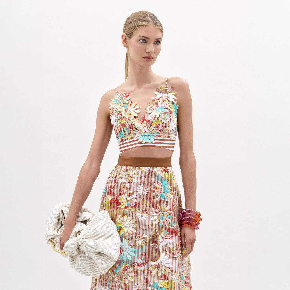 A woman wearing the Ibbie Top, a floral embroidered cropped top made from luxurious Italian fabric, pairs it with a matching skirt, holds a large textured clutch, and wears red bangles, standing against a plain white background.