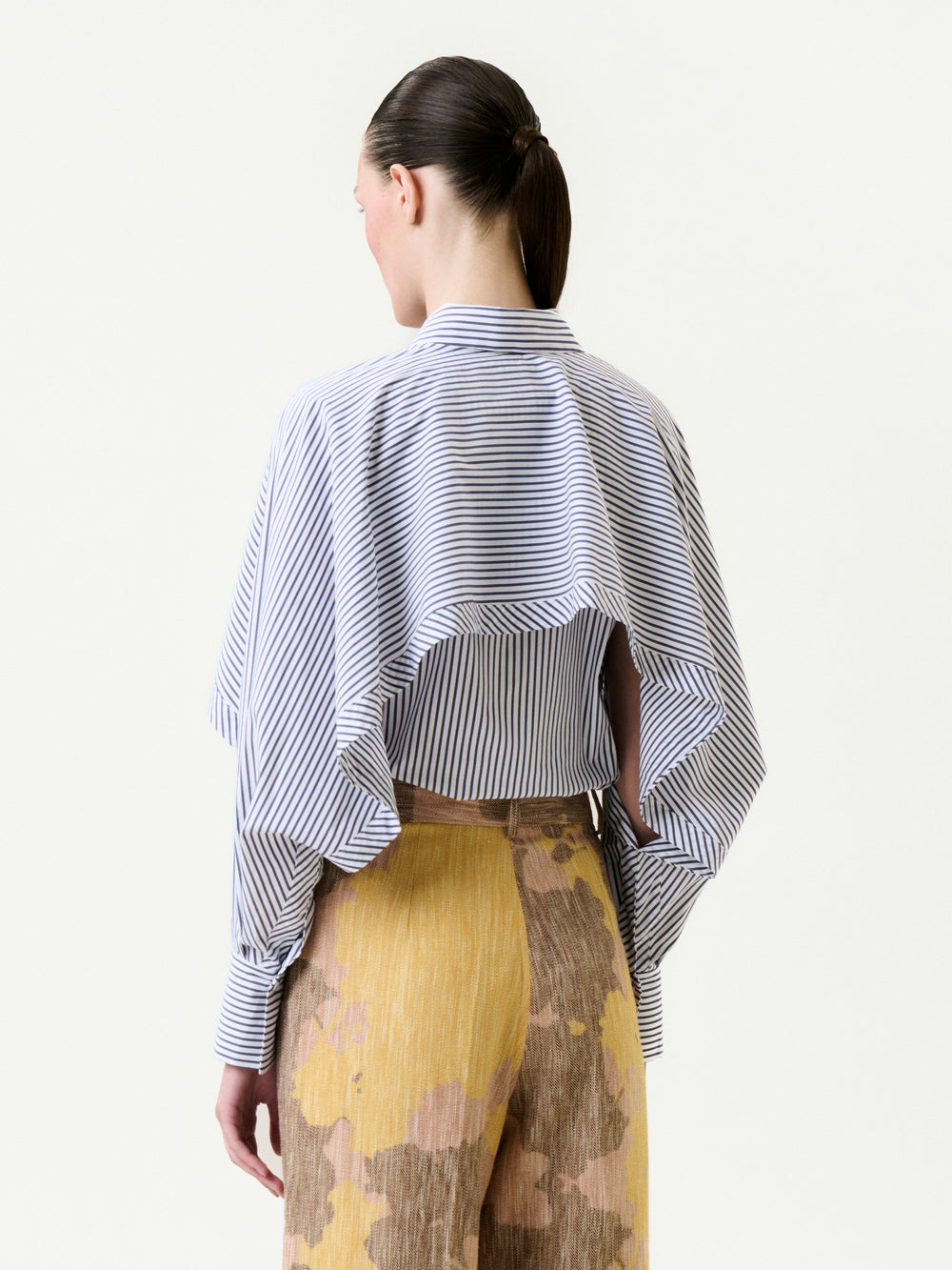 A person with dark hair in a low ponytail wears the Nira Shirt, a relaxed fit blue-and-white striped Italian fabric top with an open back and loose sleeves, paired with pleated tan pants featuring yellow and brown abstract patterns.
