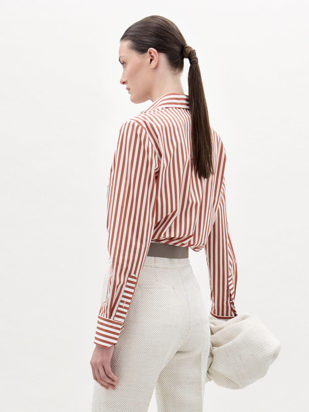 A woman stands against a plain background wearing the Karole Blouse, featuring stripes and floral embroidery, paired with textured white pants, a statement belt, and holding a folded cream-colored garment.
