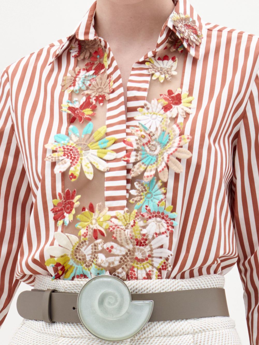 A woman stands against a plain background wearing the Karole Blouse, featuring stripes and floral embroidery, paired with textured white pants, a statement belt, and holding a folded cream-colored garment.