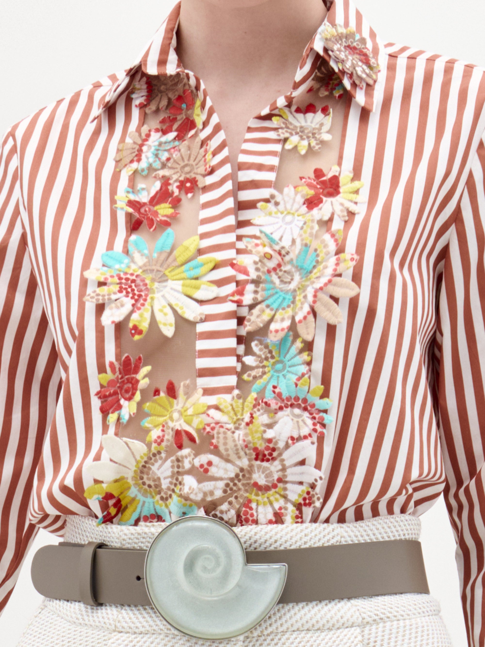 A woman stands against a plain background wearing the Karole Blouse, featuring stripes and floral embroidery, paired with textured white pants, a statement belt, and holding a folded cream-colored garment.