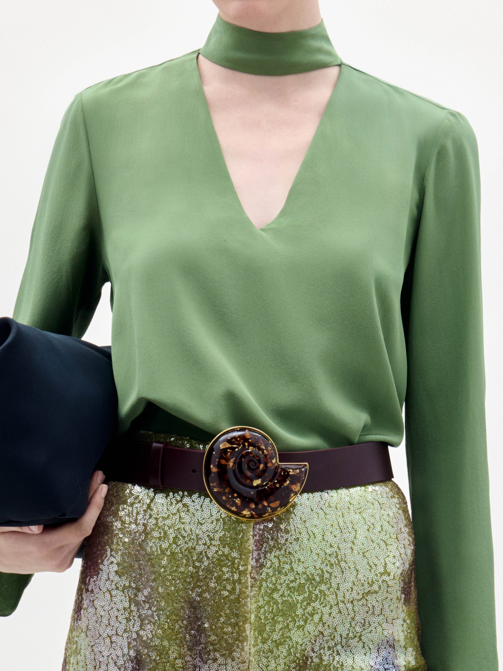 A woman poses against a plain background wearing the green Kelsey Blouse, sequined pants, a wide belt, and holding a black clutch.