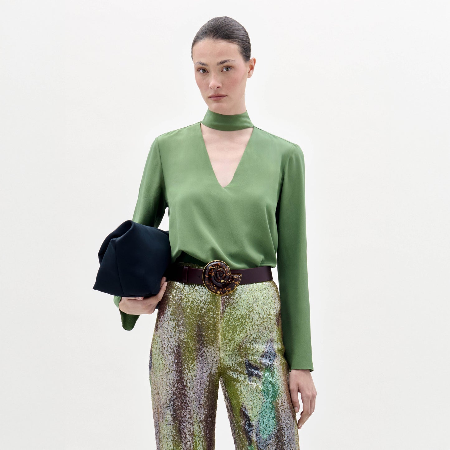 A woman poses against a plain background wearing the green Kelsey Blouse, sequined pants, a wide belt, and holding a black clutch.