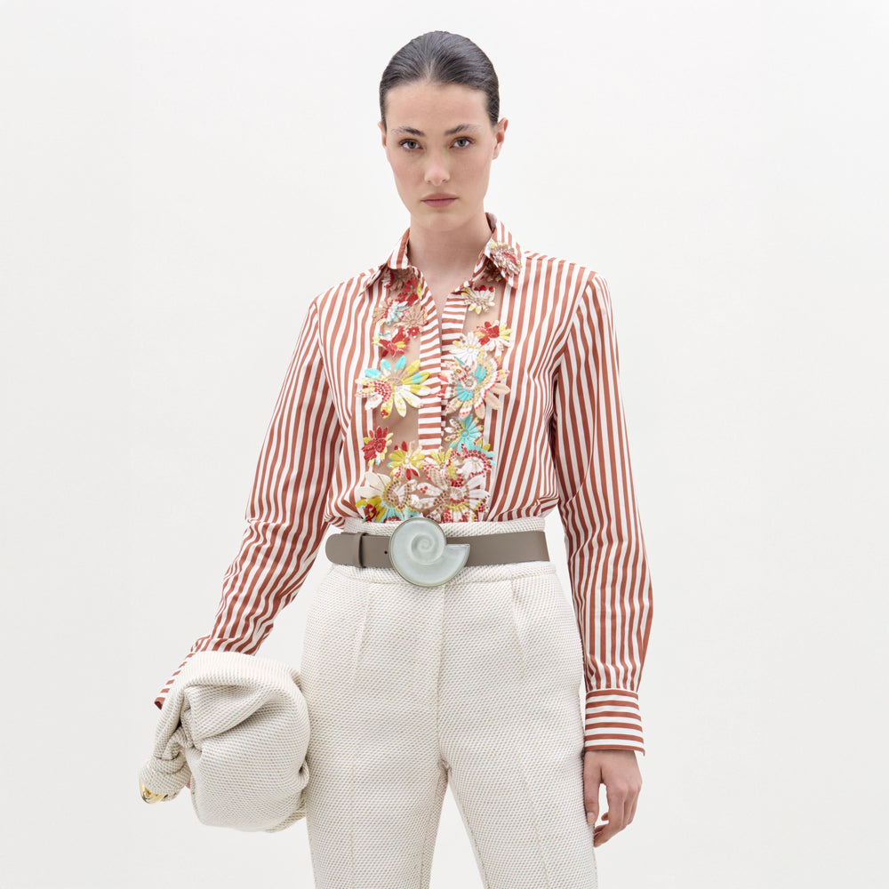 A woman stands against a plain background wearing the Karole Blouse, featuring stripes and floral embroidery, paired with textured white pants, a statement belt, and holding a folded cream-colored garment.