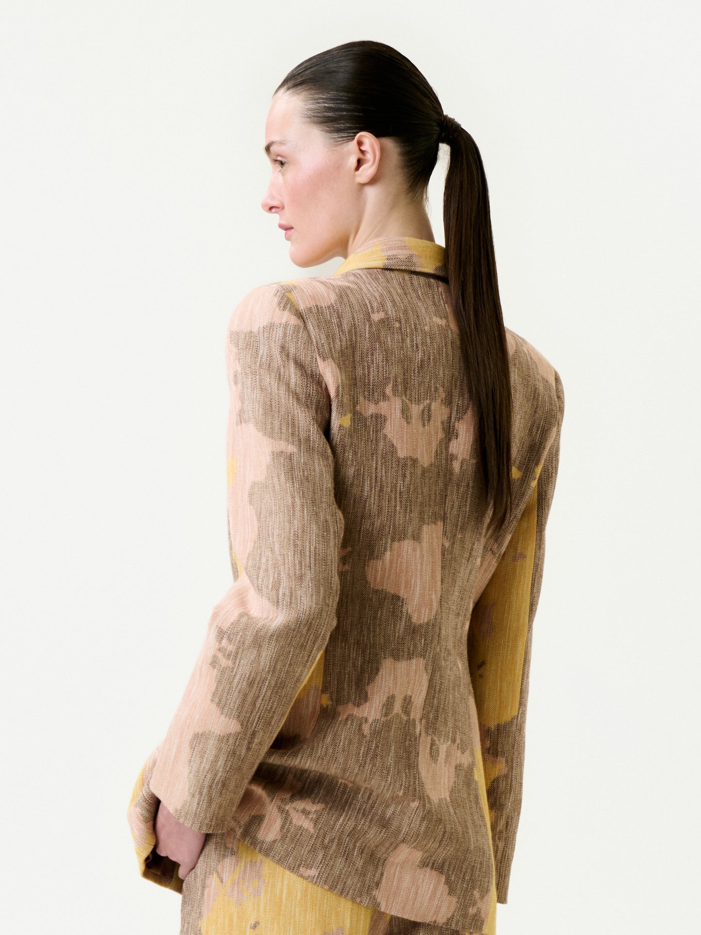 A woman with straight, dark hair in a low ponytail models the Frankie Jacket, an Italian fabric blazer in light brown and beige pattern with a true-to-size fit, as she looks to the side against a plain white background.