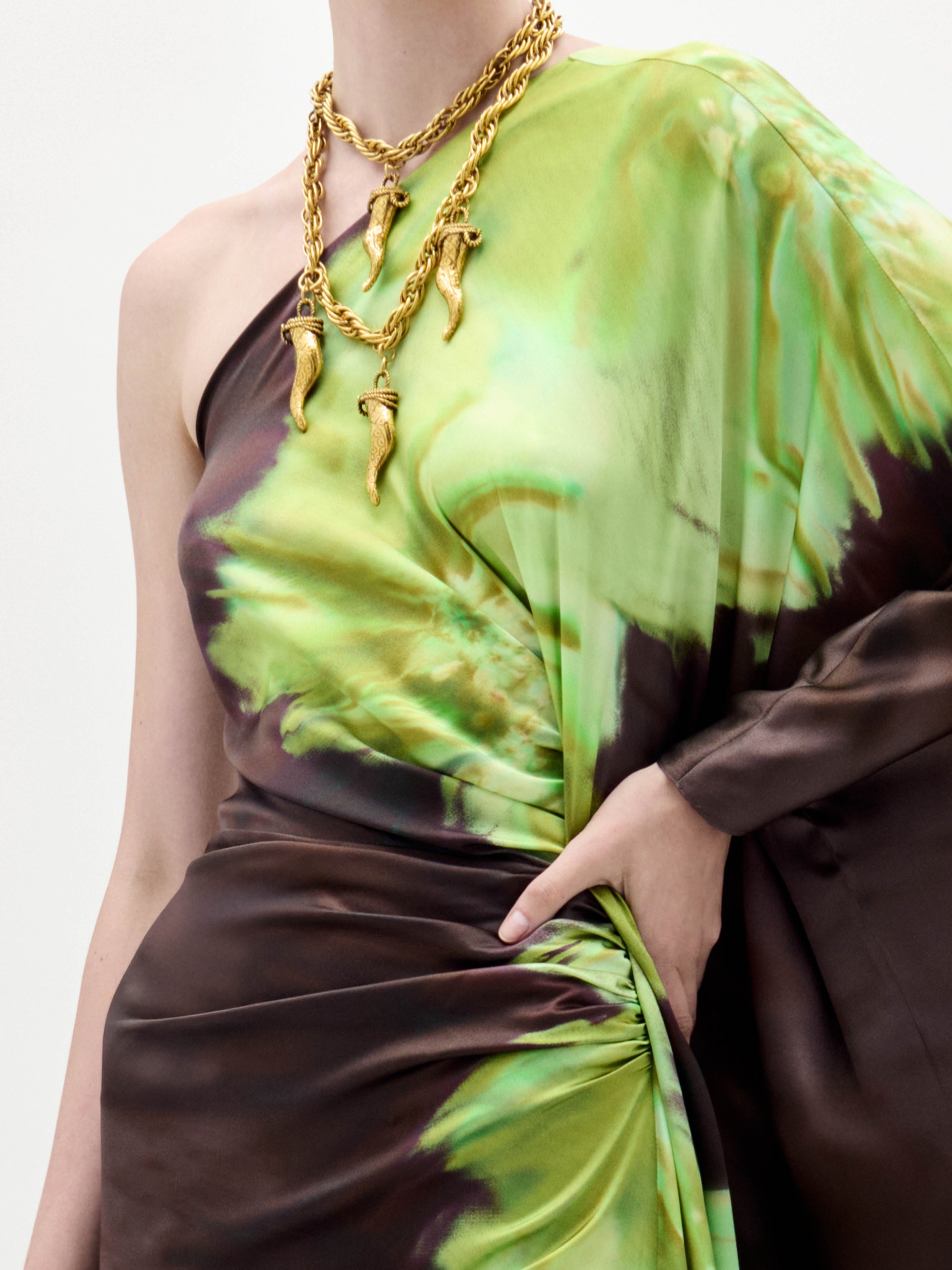 A person models the Afrodita Dress—a one-shoulder, floor-length gown with a green and brown tie-dye print—paired with gold necklaces, against a plain white background.