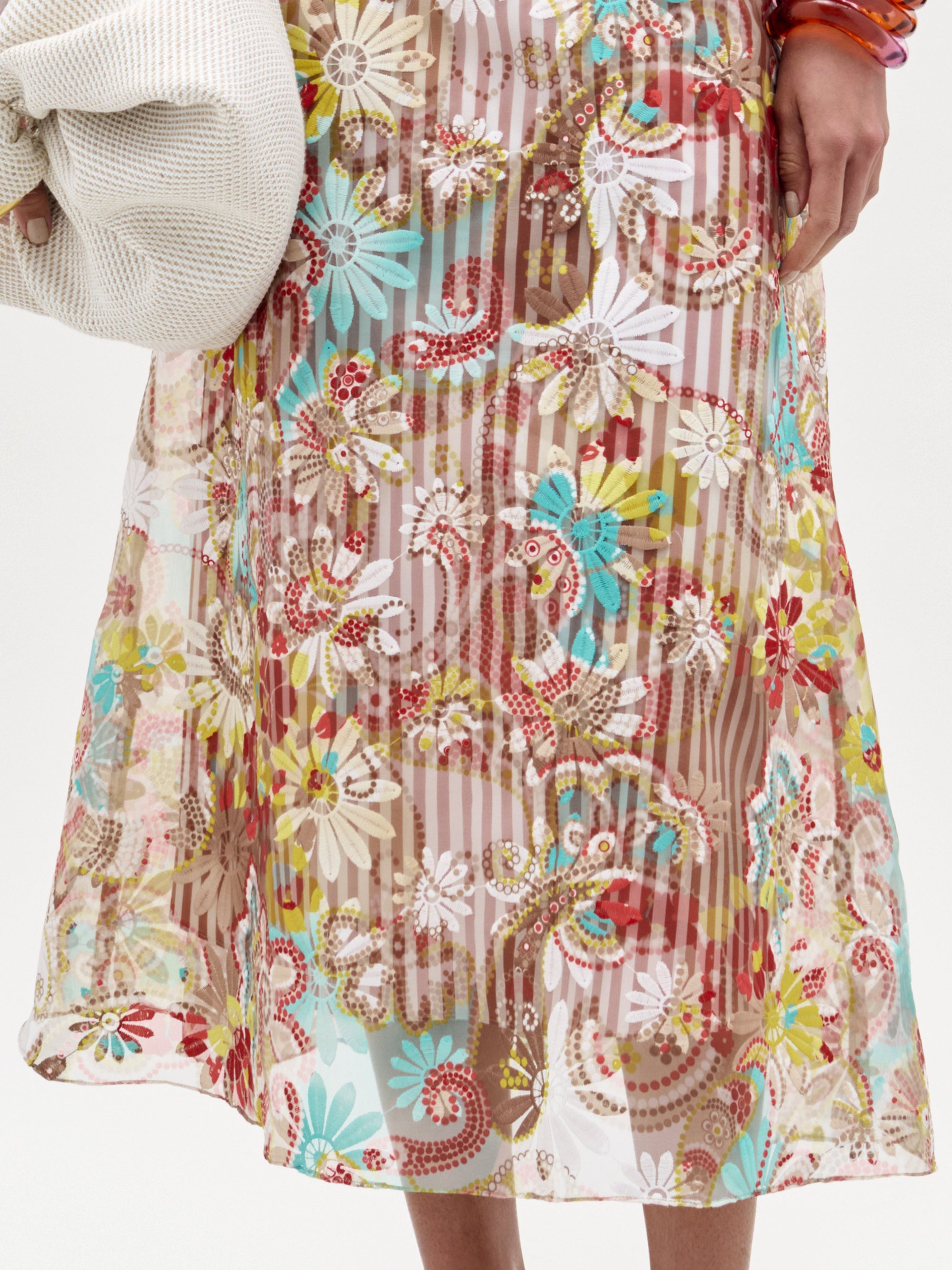 A woman stands against a plain white background, wearing the Nelida Skirt—a floral, sheer, A-line midi skirt with a matching long-sleeved blouse—paired with heeled sandals and a large white clutch.