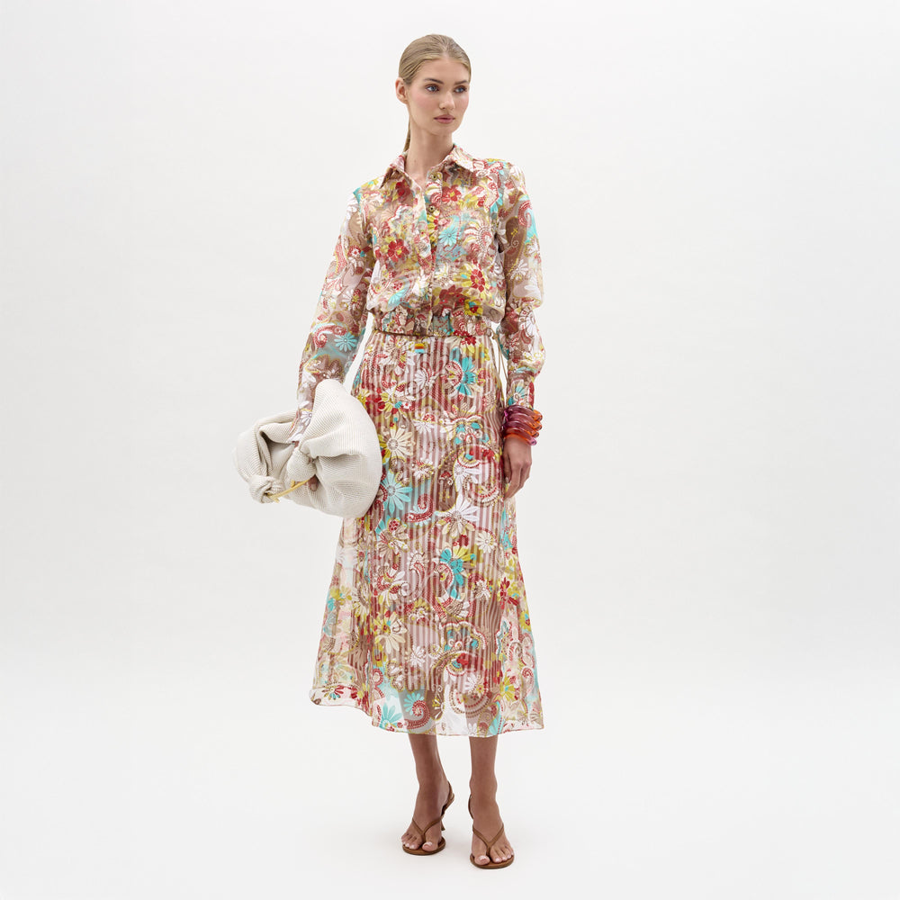 A woman stands against a plain white background, wearing the Nelida Skirt—a floral, sheer, A-line midi skirt with a matching long-sleeved blouse—paired with heeled sandals and a large white clutch.