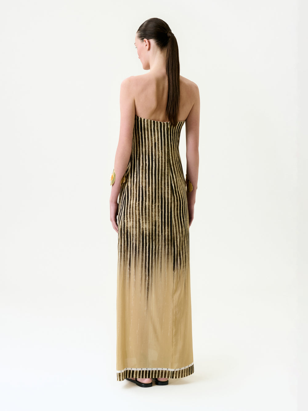 A person with long brown hair in a low ponytail faces away from the camera, wearing the Lirit Dress—a strapless, floor-length maxi crafted from Italian fabric with vertical black stripes on a beige background—against a plain white backdrop.
