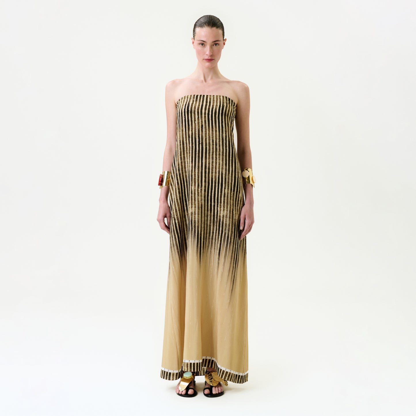 A woman faces forward against a plain white background, wearing the Lirit Dress—a strapless maxi made from luxurious Italian fabric with vertical black stripes fading into beige—paired with bracelets and black sandals. Her hair is pulled back and her expression is minimal.