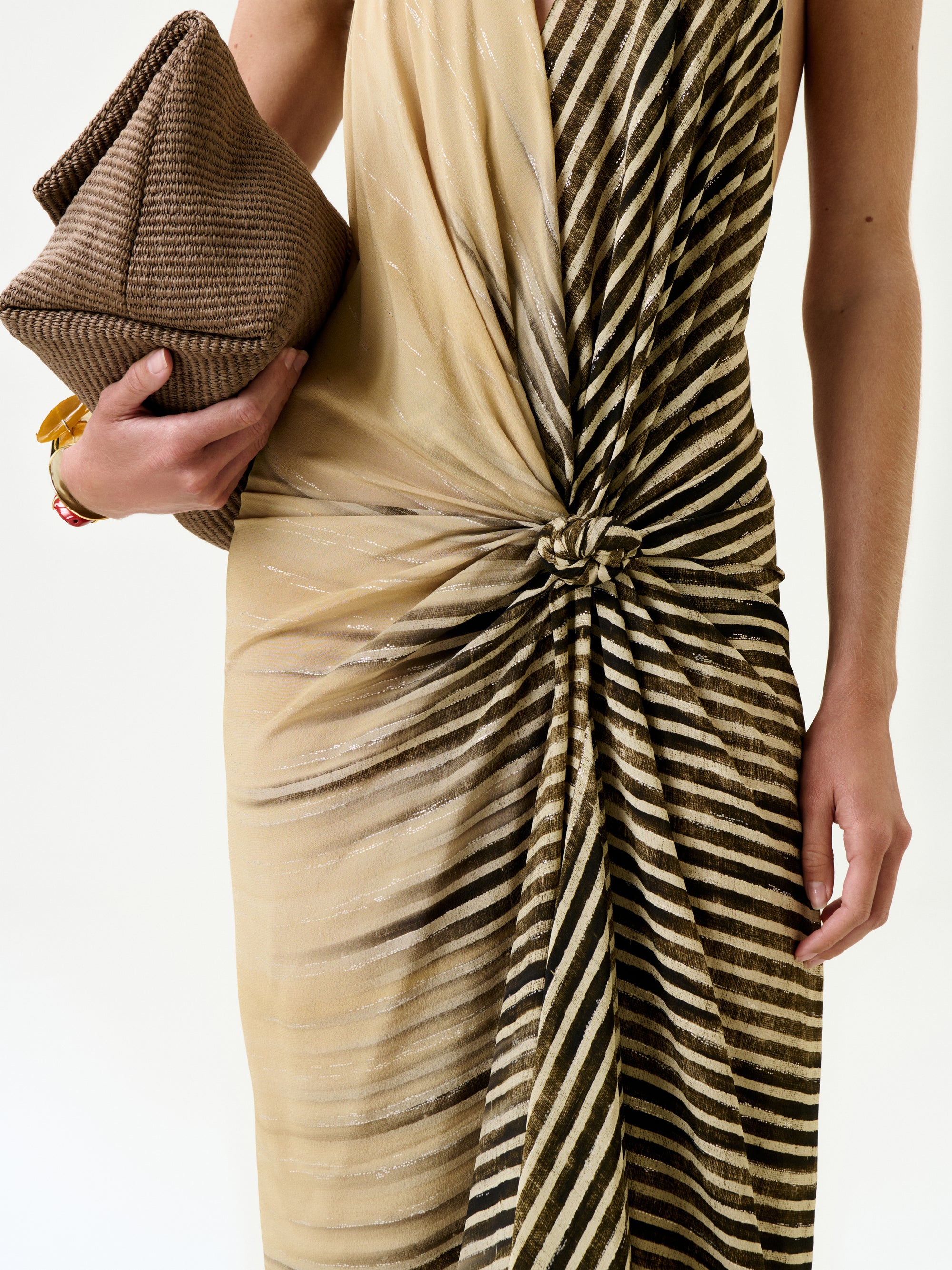 A person wears the Lorde Dress—a sleeveless beige and brown striped midi with a knotted front, made from Italian fabric—holding a large textured brown clutch. Only their torso and arms are visible against a plain background.
