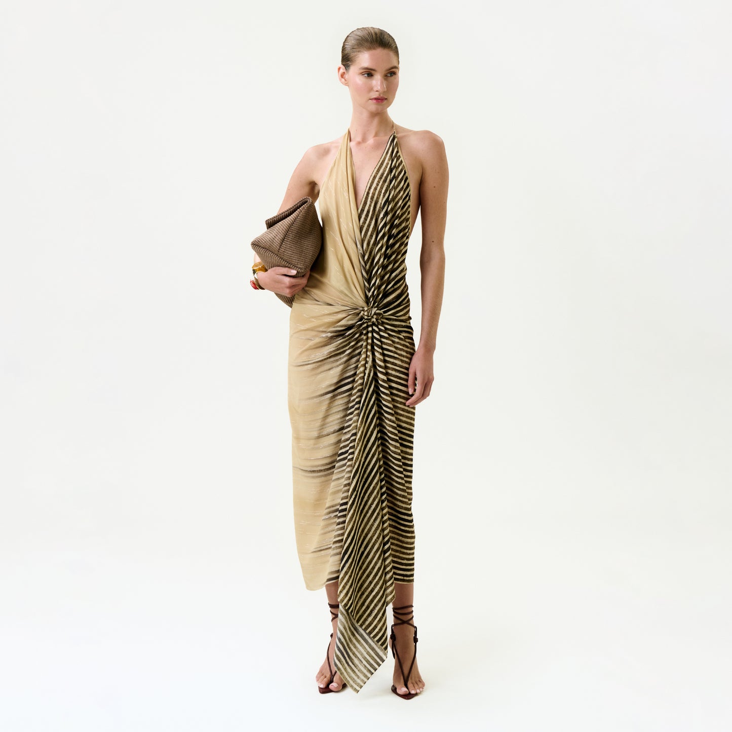 A woman stands against a plain background, wearing the Lorde Dress—a beige and black striped silk midi made from Italian fabric. She pairs it with strappy heeled sandals, gold bracelets, slicked-back hair, and a brown clutch.