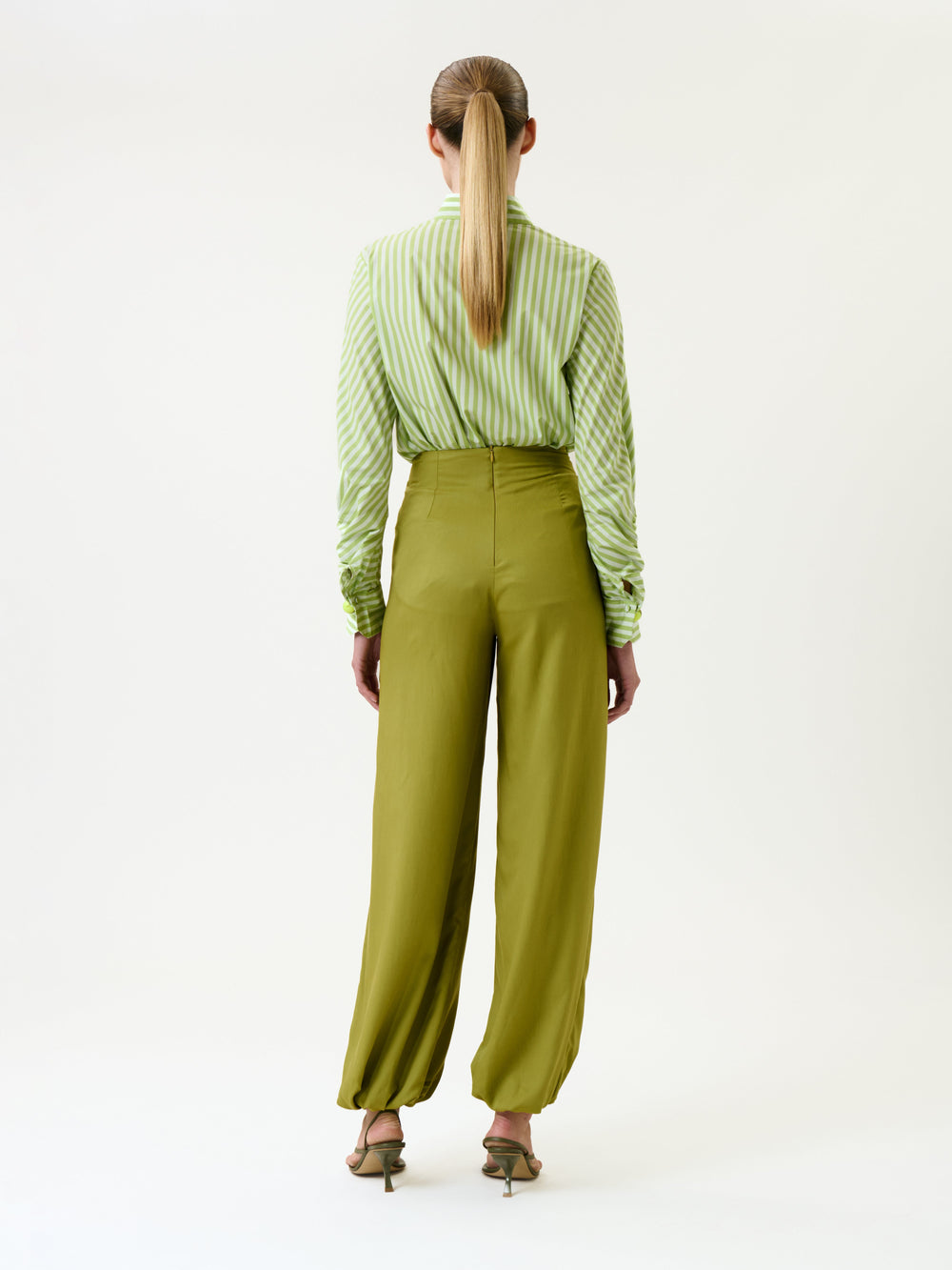 A woman stands with her back to the camera, wearing the Raimondo Pant—high-waisted, wide-leg olive trousers—paired with a green and white striped shirt and heeled sandals. Her hair is styled in a low ponytail, and she is set against a plain white background.