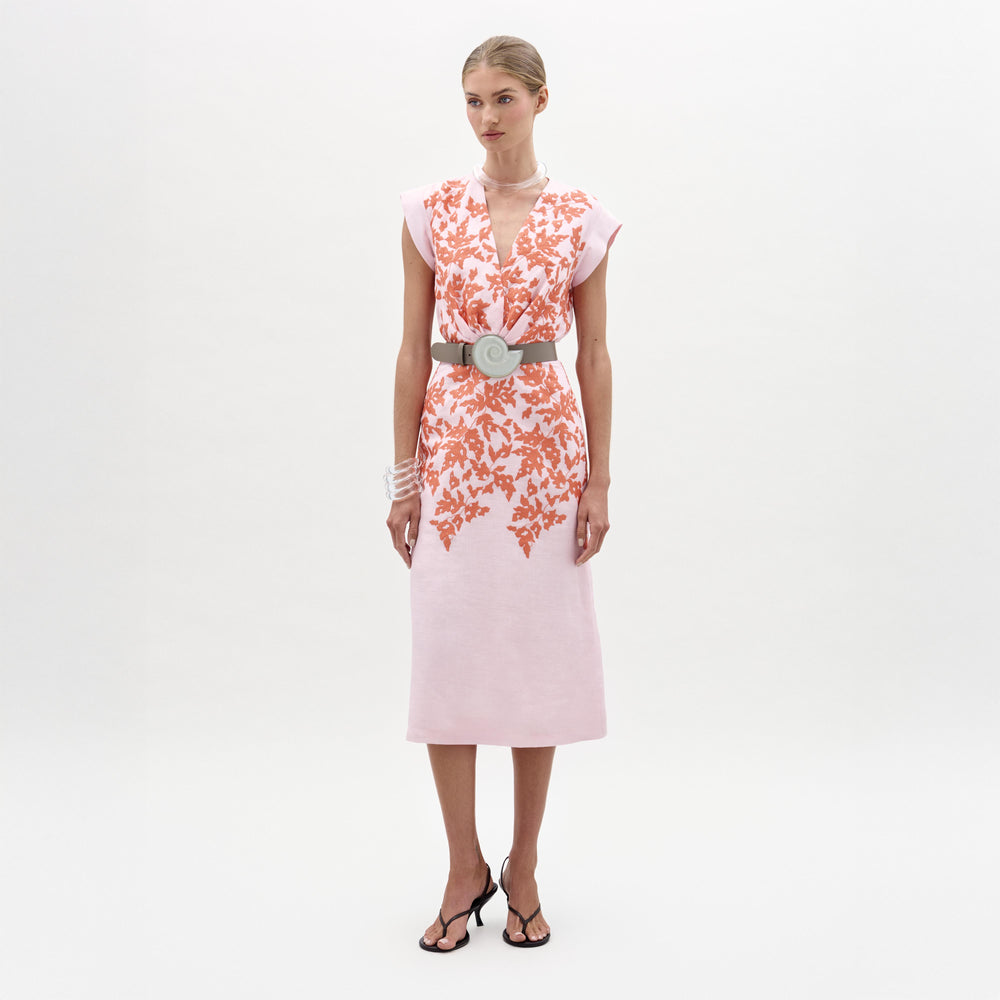 A woman stands against a plain background wearing the Olive Dress—a light pink midi dress with orange floral patterns, a fitted waist accented by a wide gray belt, and black heeled sandals.