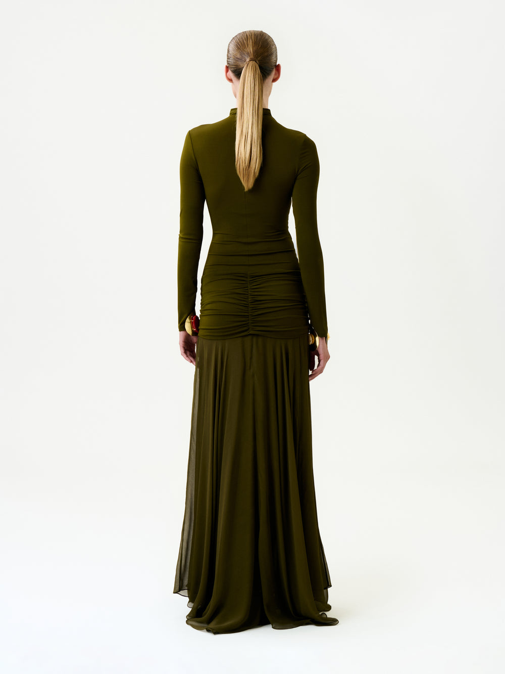 A person with long, straight hair in a low ponytail is shown from the back wearing the Monie Dress, a long-sleeved, floor-length olive green maxi dress, against a plain white background.