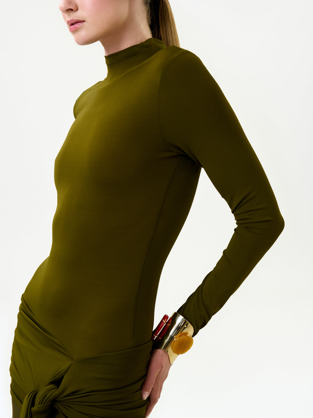 A person wears the Monie Dress—a fitted, long-sleeve olive green maxi dress with a high neckline and side knot—paired with chunky bracelets and tied-back hair against a plain white background.