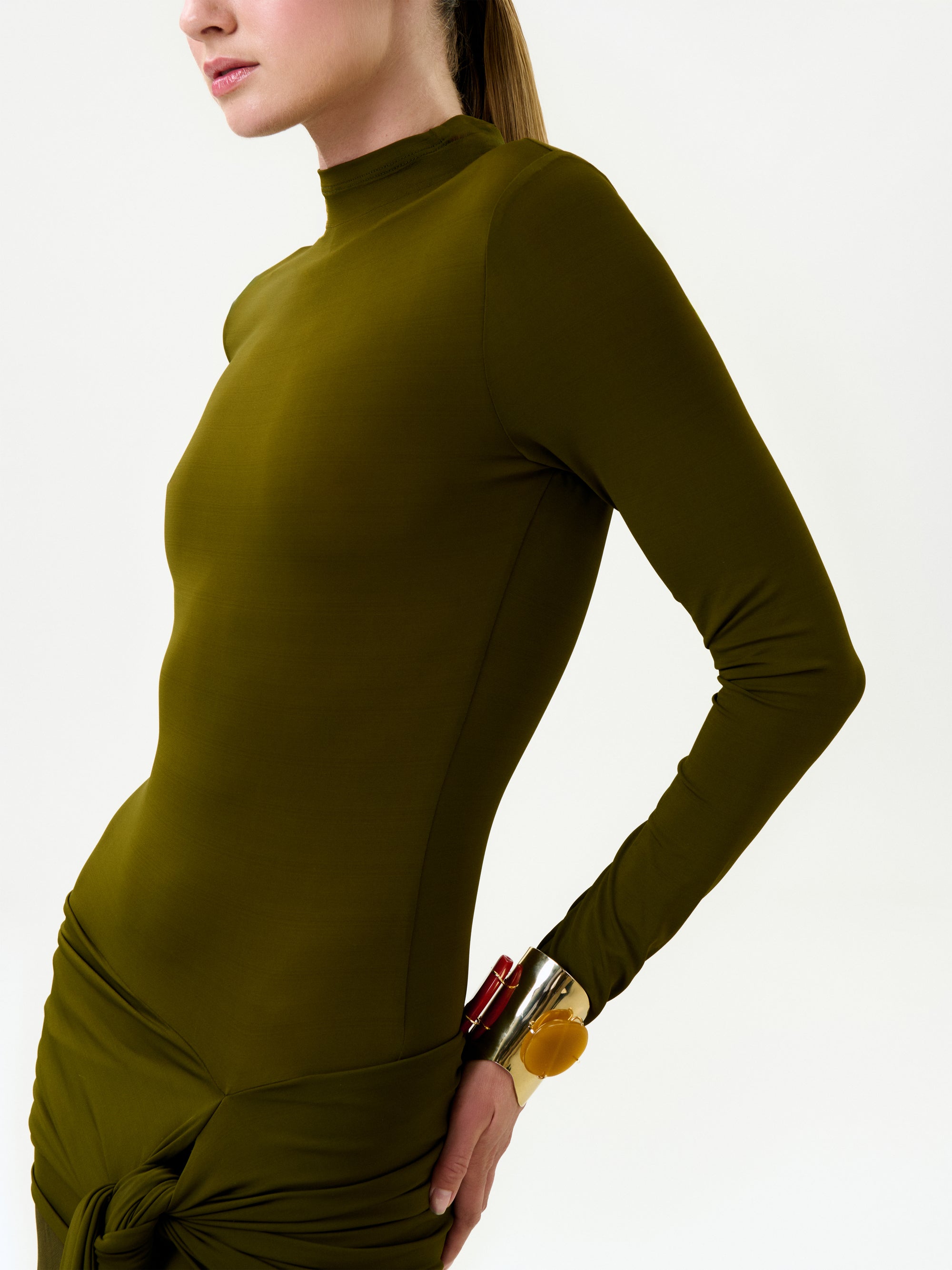 A person wears the Monie Dress—a fitted, long-sleeve olive green maxi dress with a high neckline and side knot—paired with chunky bracelets and tied-back hair against a plain white background.