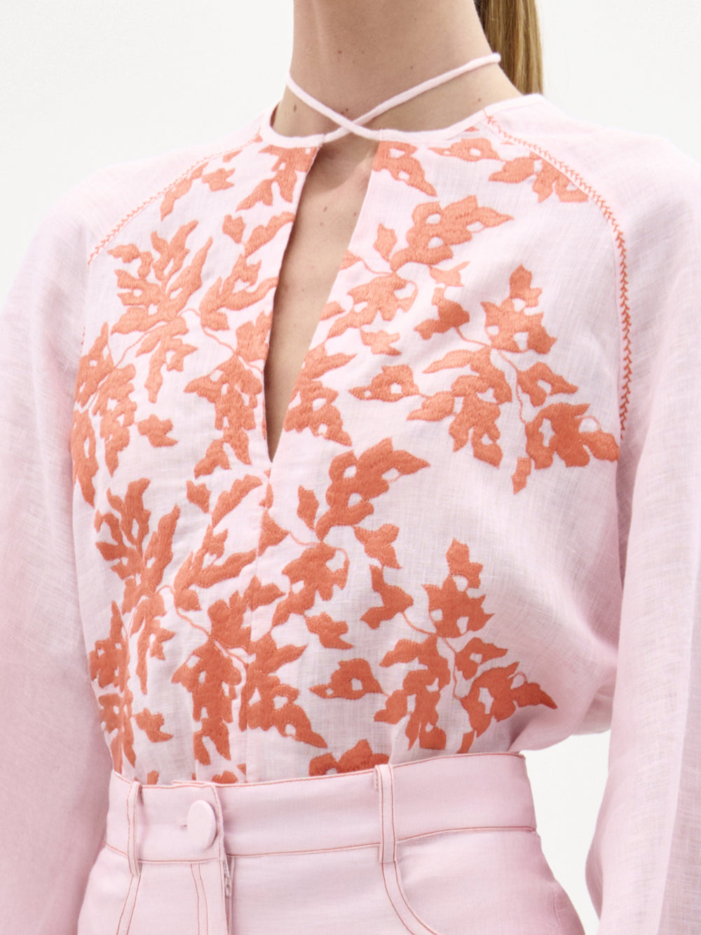 A woman stands against a plain background wearing the Molveno Blouse with a V neckline, a matching skirt, and holding a pale pink clutch.