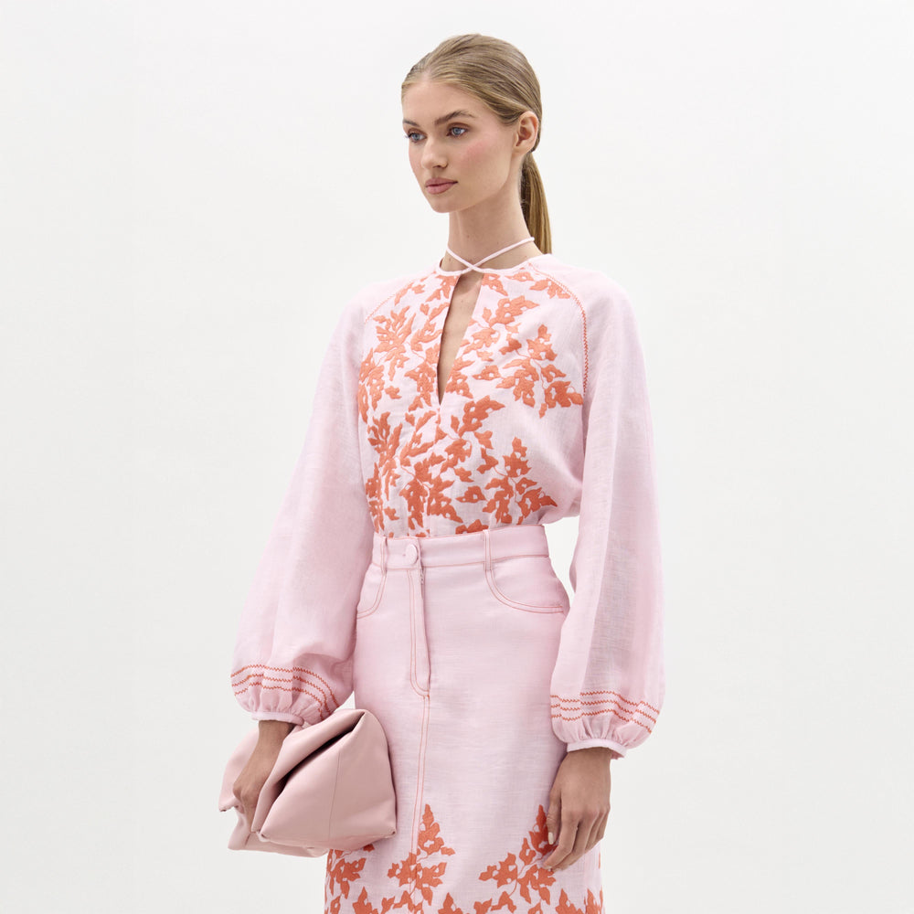 A woman stands against a plain background wearing the Molveno Blouse with a V neckline, a matching skirt, and holding a pale pink clutch.