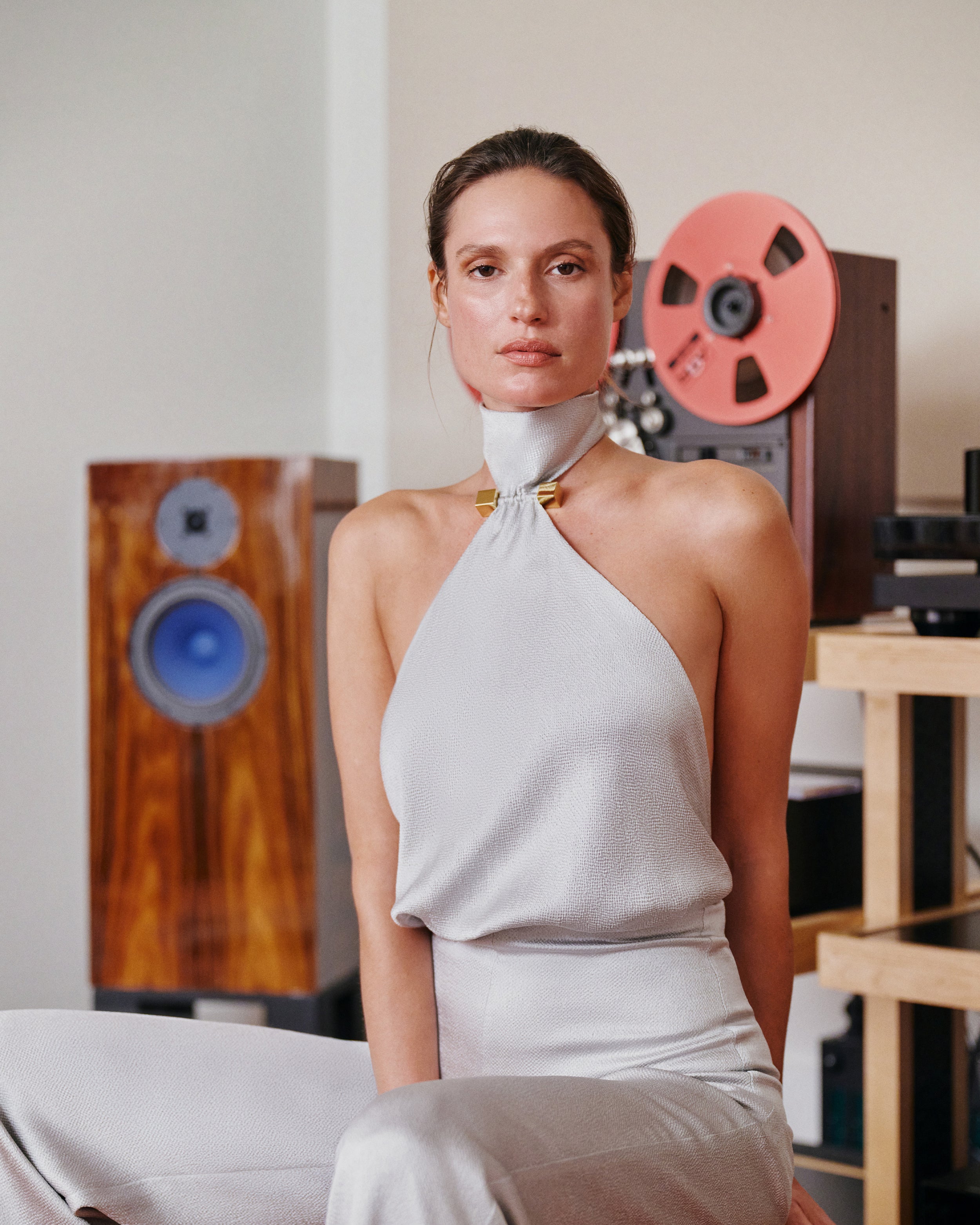 A woman wearing a light gray halter-neck top and matching pants sits in a room with vintage audio equipment, including speakers and a reel-to-reel recorder, in the background.