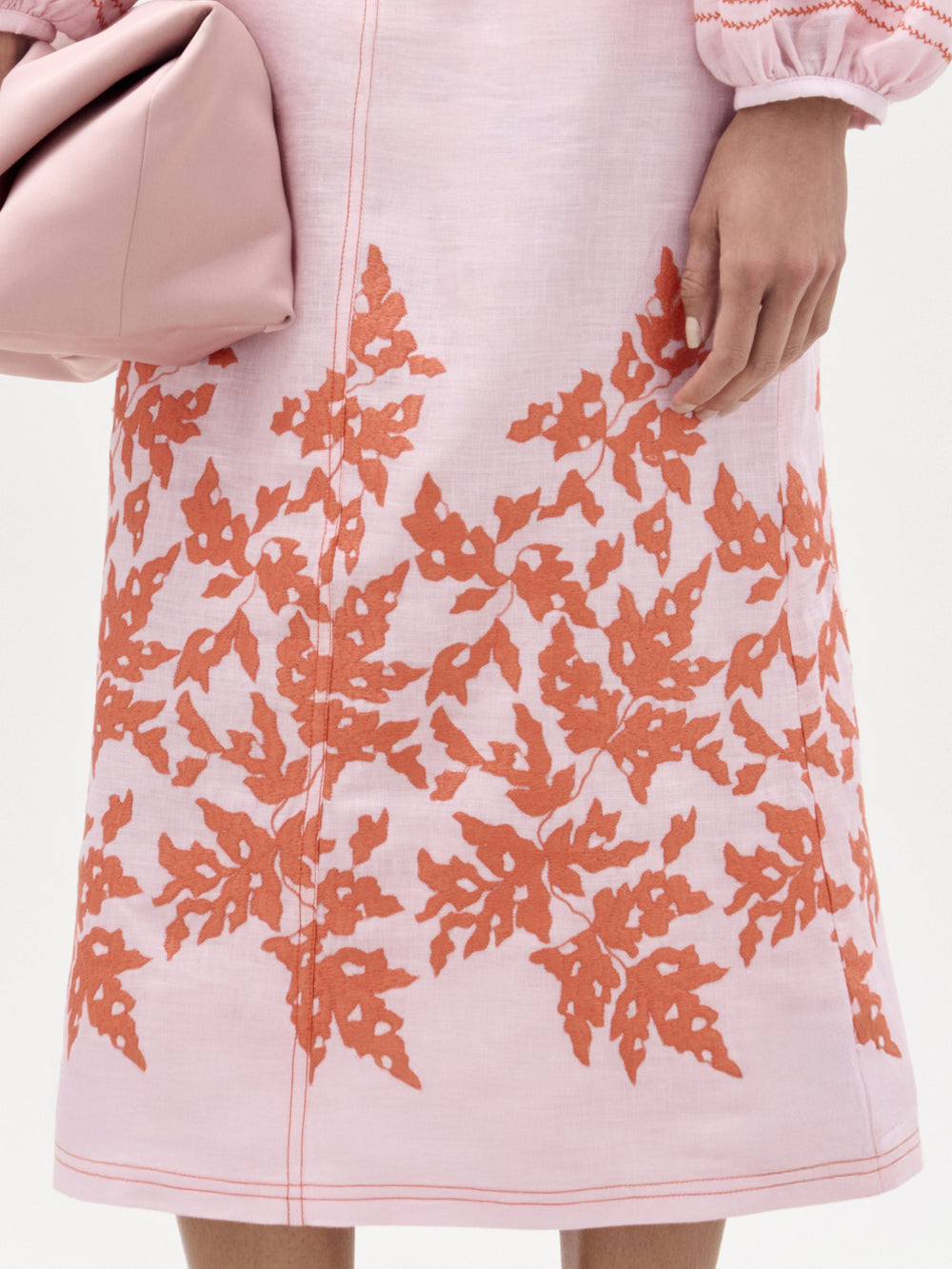 A woman stands against a plain background wearing a pink blouse, the Layan Skirt—a structured midi skirt with orange floral embroidery—paired with heeled sandals and holding a pink clutch.