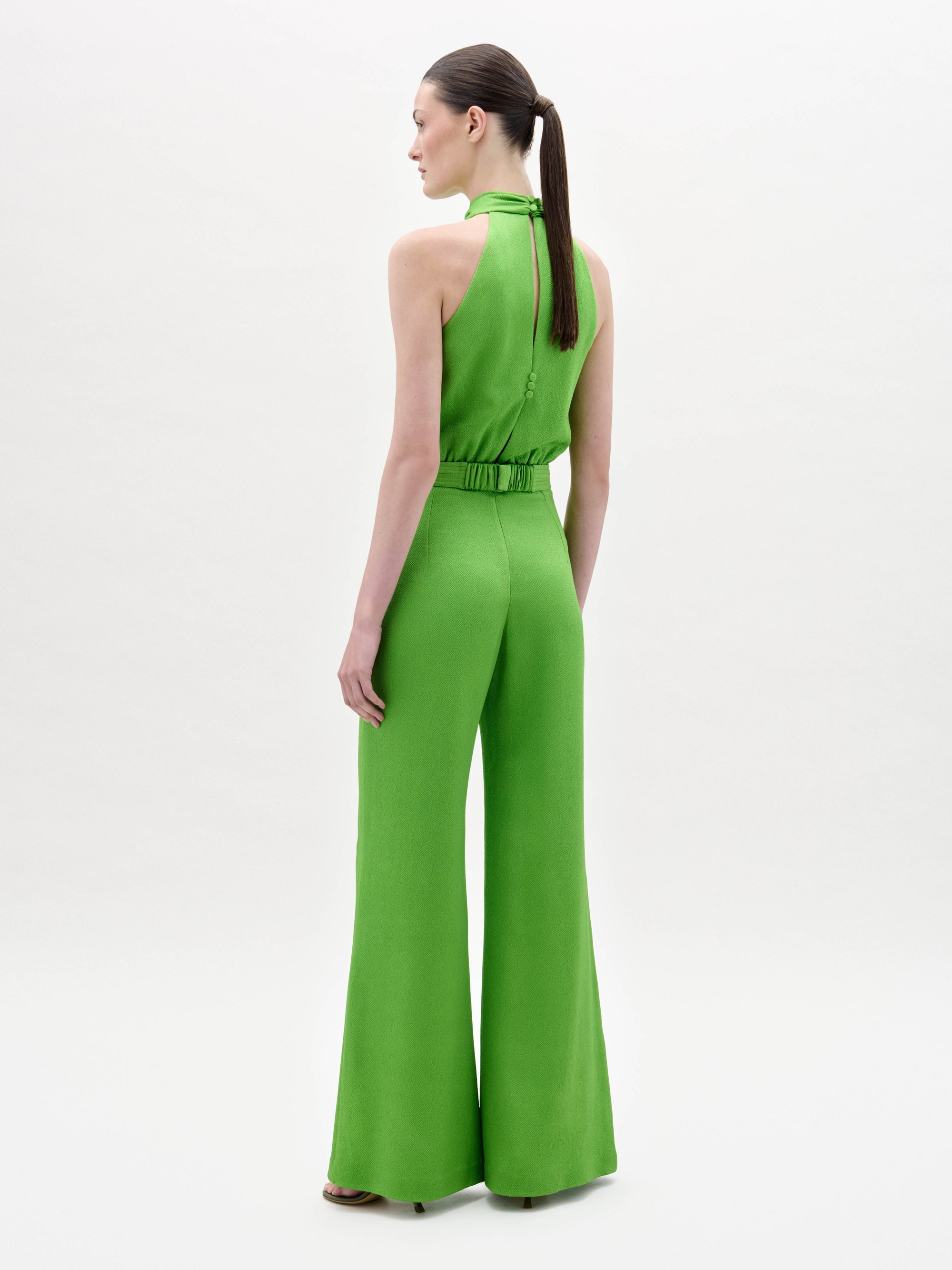 A woman stands facing forward in the Siv Jumpsuit, a bright green halter-neck style with wide-leg pants and a matching belt, paired with black sandals, against a plain white background.