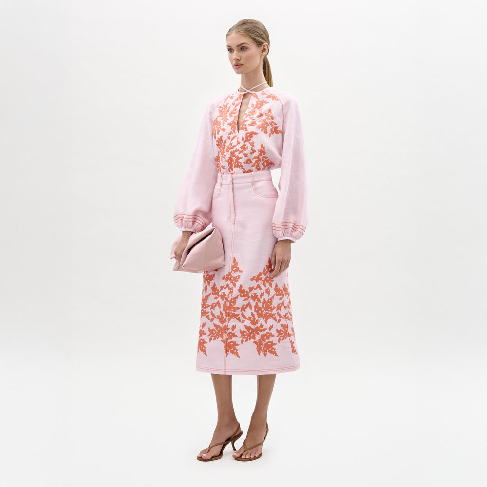 A woman stands against a plain background wearing a pink blouse, the Layan Skirt—a structured midi skirt with orange floral embroidery—paired with heeled sandals and holding a pink clutch.