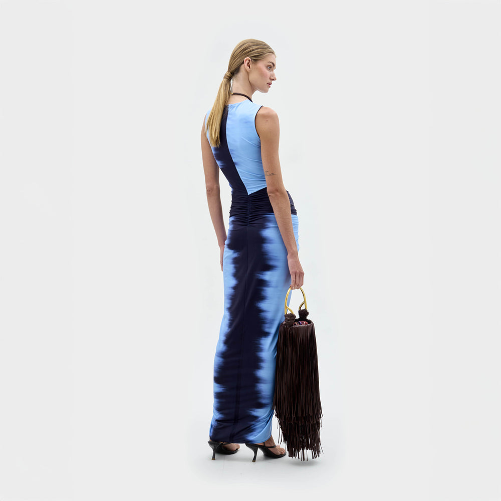 A woman wears the Leni Dress—a sleeveless blue satin dress with gathered detail and front slit—paired with a large black fringe handbag and black heeled sandals, standing against a plain background.