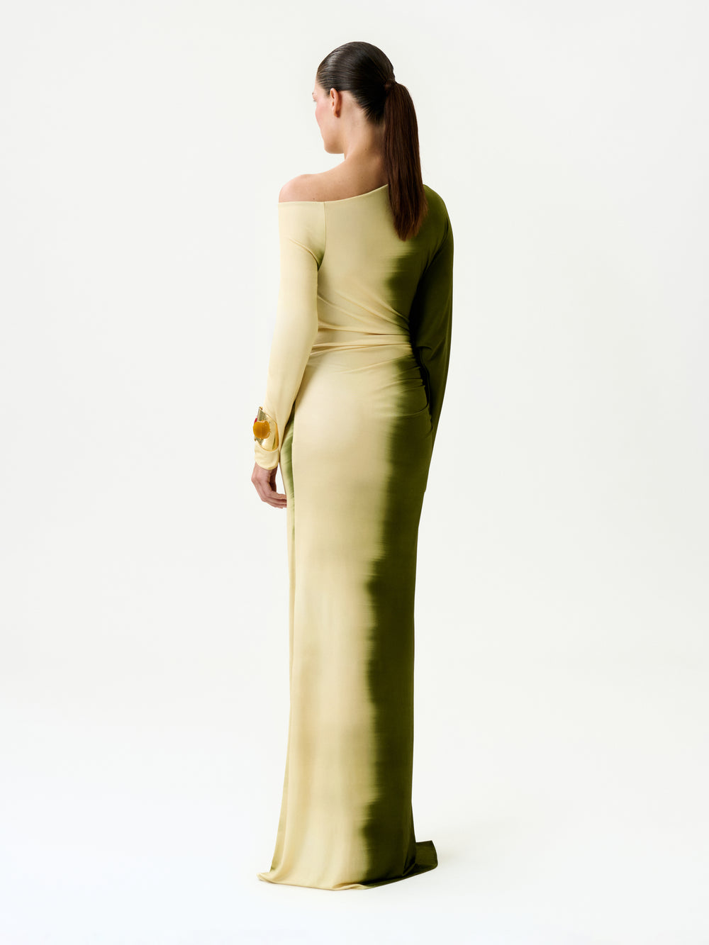 A person with long brown hair in a ponytail stands with their back to the camera against a white background, wearing the Nadja Dress—a form-fitting, off-shoulder ombré yellow and green maxi dress made in Colombia.