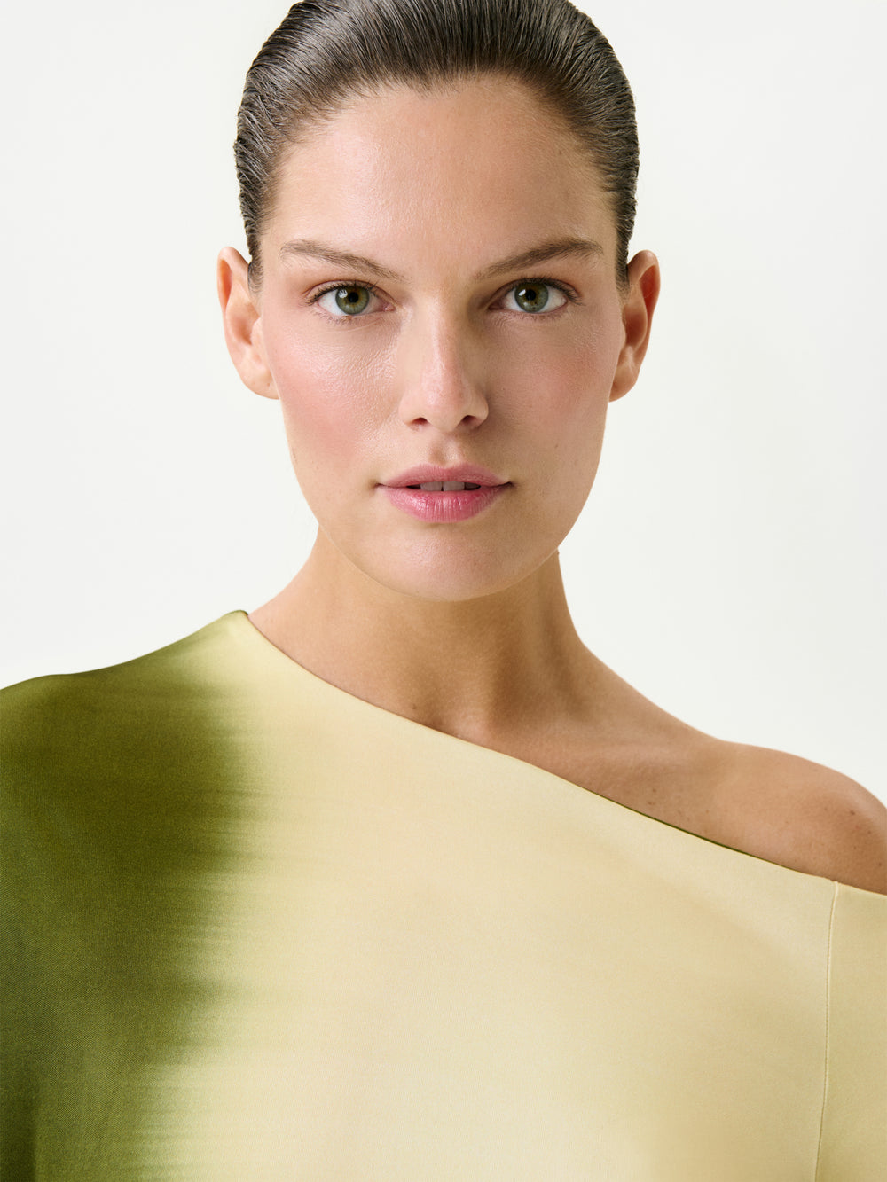 A woman with slicked-back brown hair and green eyes wears the Nadja Dress—an off-the-shoulder maxi in a green-to-cream gradient, crafted from fine Italian fabric—posing against a plain white background.