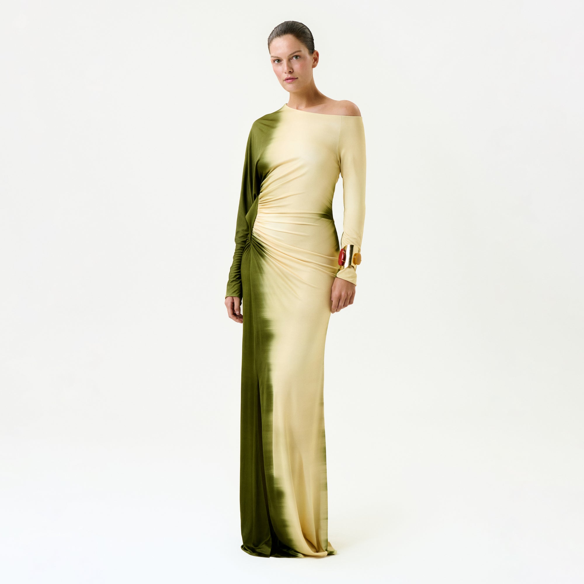 A person stands against a white background wearing the Nadja Dress—a Colombian-made maxi with a gradient from olive green to pale yellow, featuring one long sleeve and an off-the-shoulder neckline.