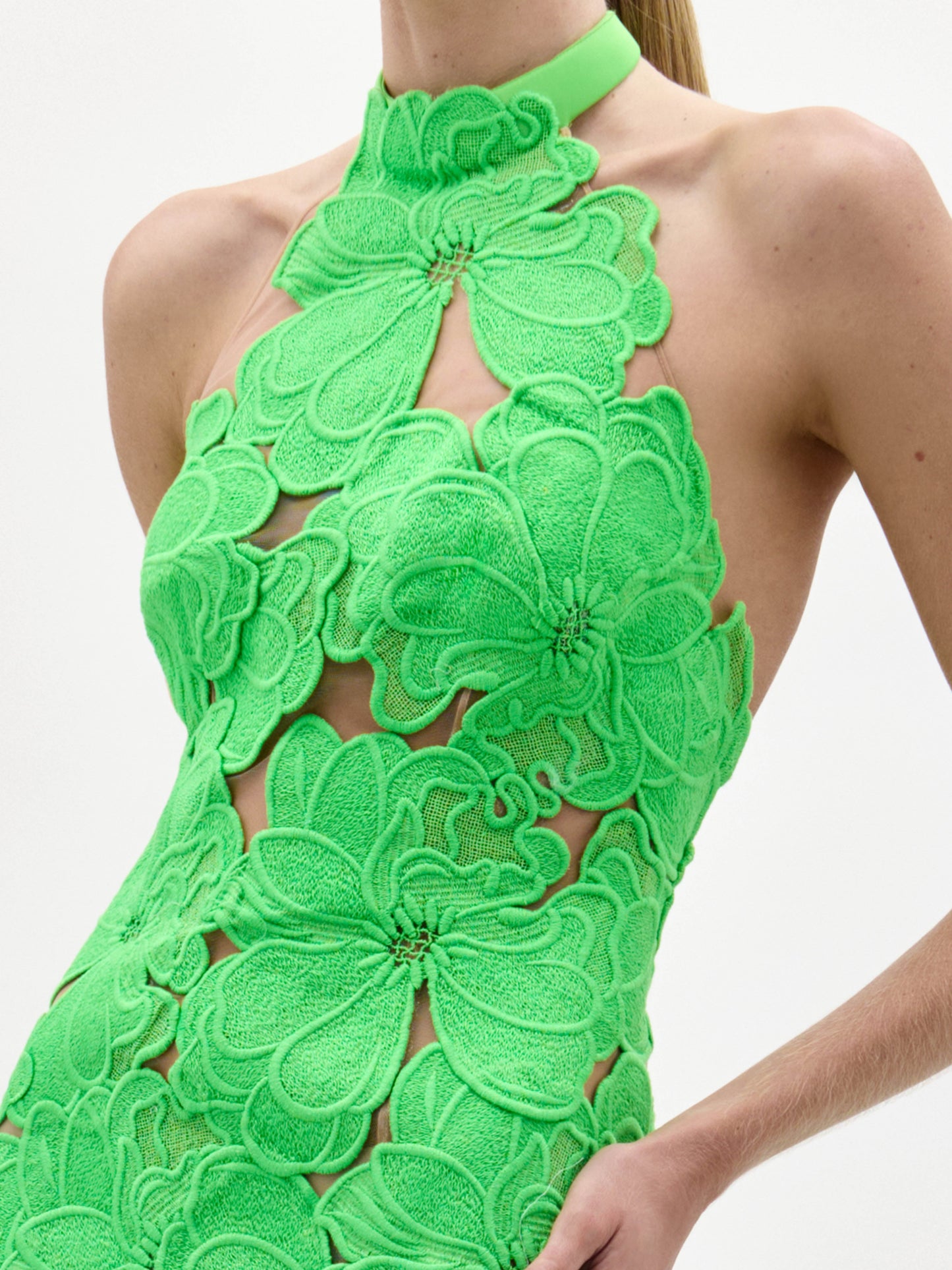A woman models the Mab Dress, a sleeveless, floor-length gown featuring large green floral appliqués on a sheer nude base, set against a plain white background.