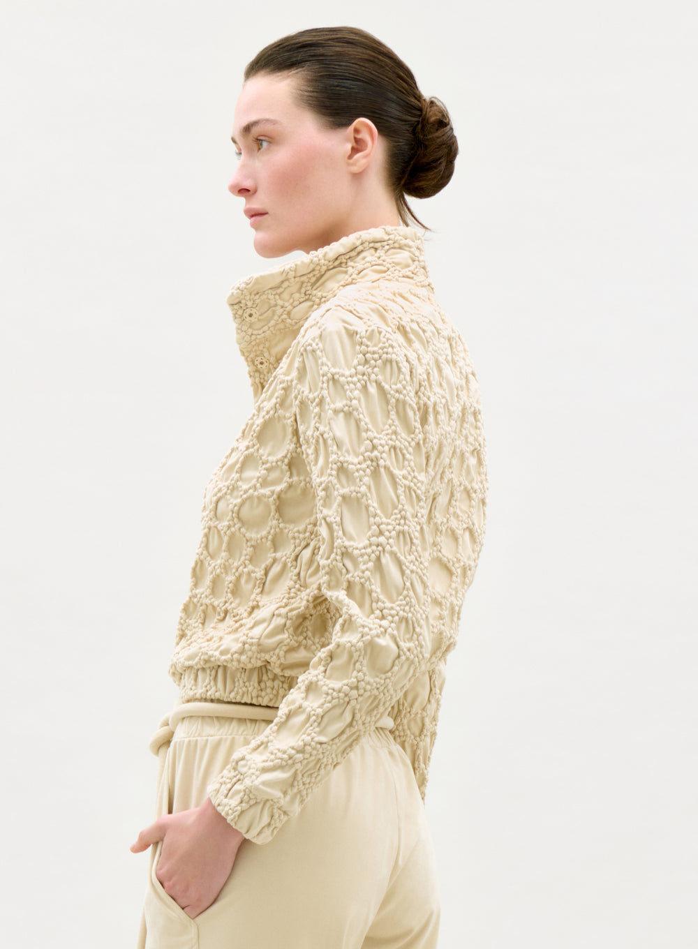 A woman with dark hair in a low bun wears the Zalta Jacket, a high-collared, textured beige piece with matching pants, standing in profile against a plain light background.