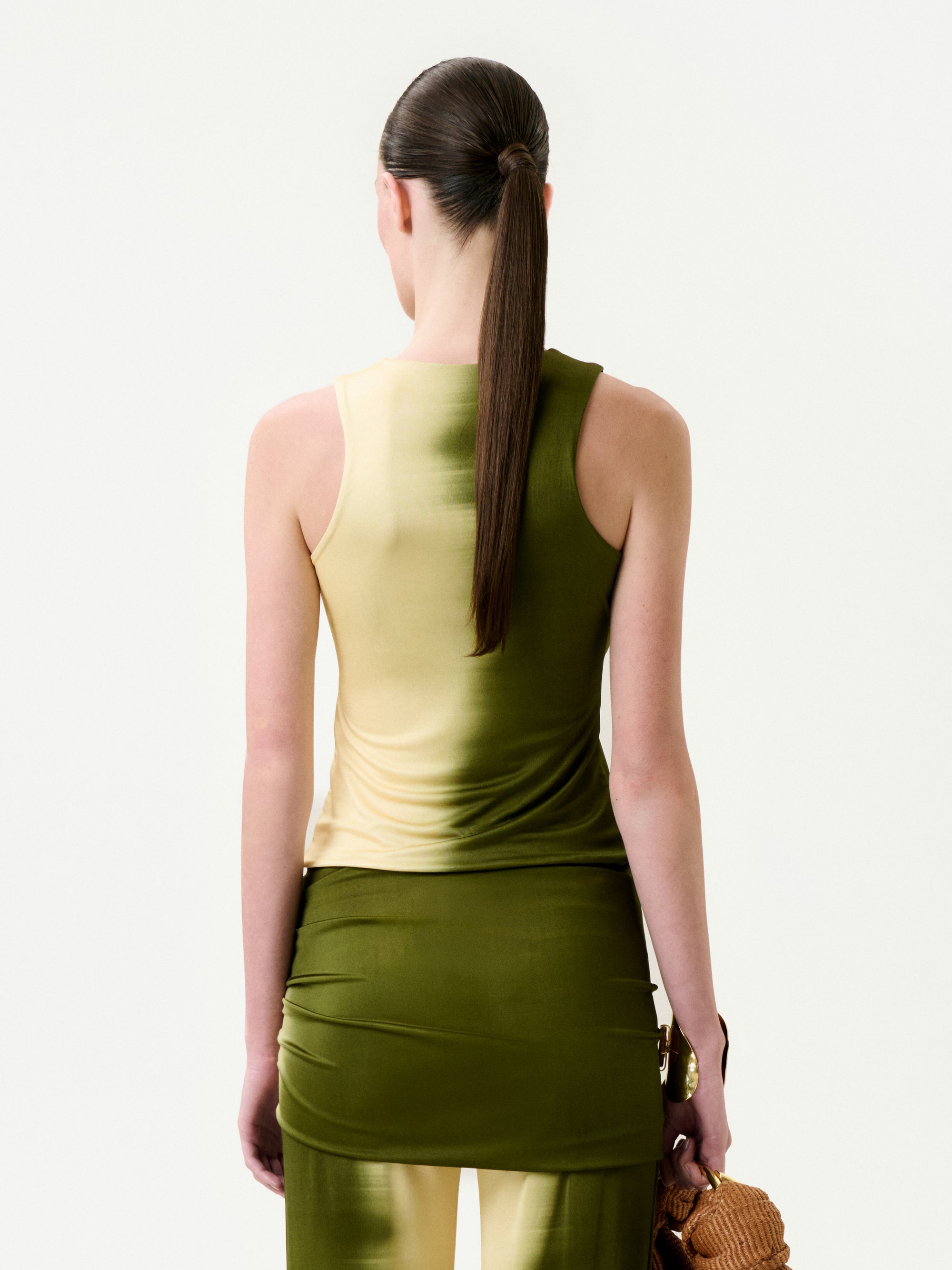 A woman with long brown hair in a low ponytail stands with her back to the camera, wearing a slim fit Tagua T-Shirt in green and beige gradient and matching skirt, while holding a brown woven handbag.