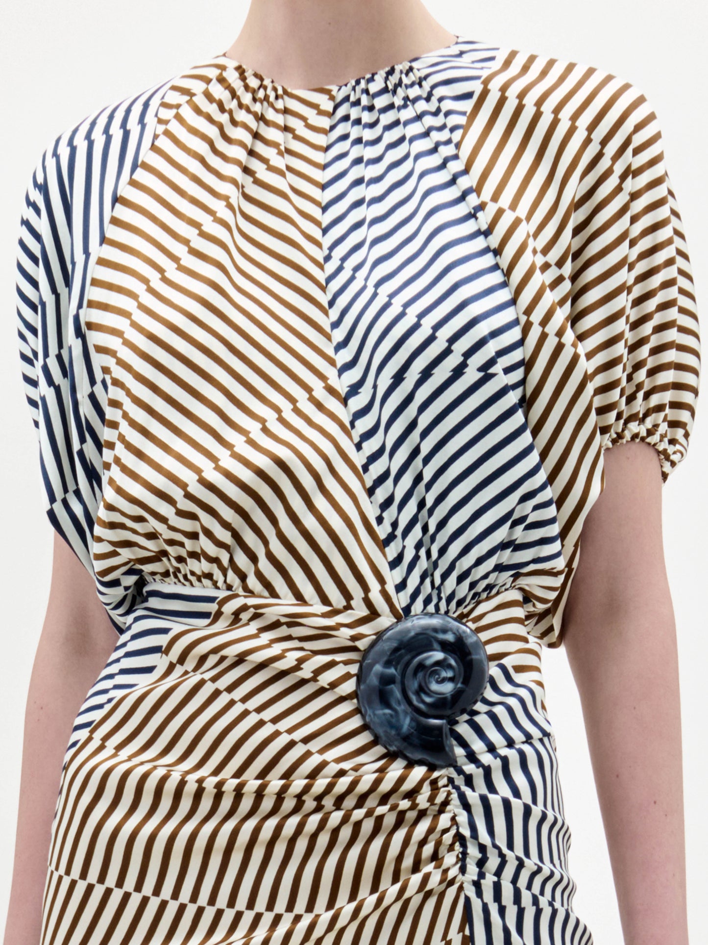 A woman models the Kala Dress, a beige and blue striped dress with a cinched waist and floral detail, holding a long fringed handbag against a plain white background.
