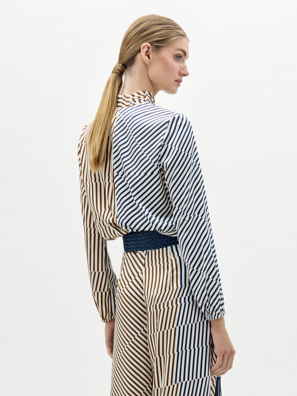 Woman wearing the Jasper Blouse, a long-sleeved, blue and beige striped top, paired with a matching choker and a wide belt with a large gold buckle, stands against a white background.
