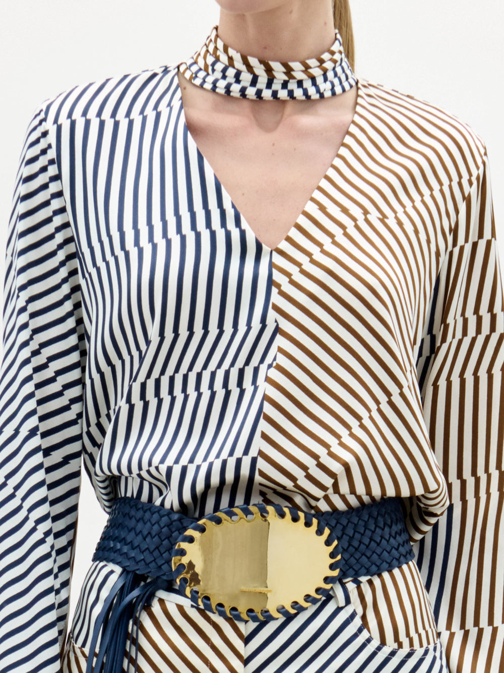 Woman wearing the Jasper Blouse, a long-sleeved, blue and beige striped top, paired with a matching choker and a wide belt with a large gold buckle, stands against a white background.